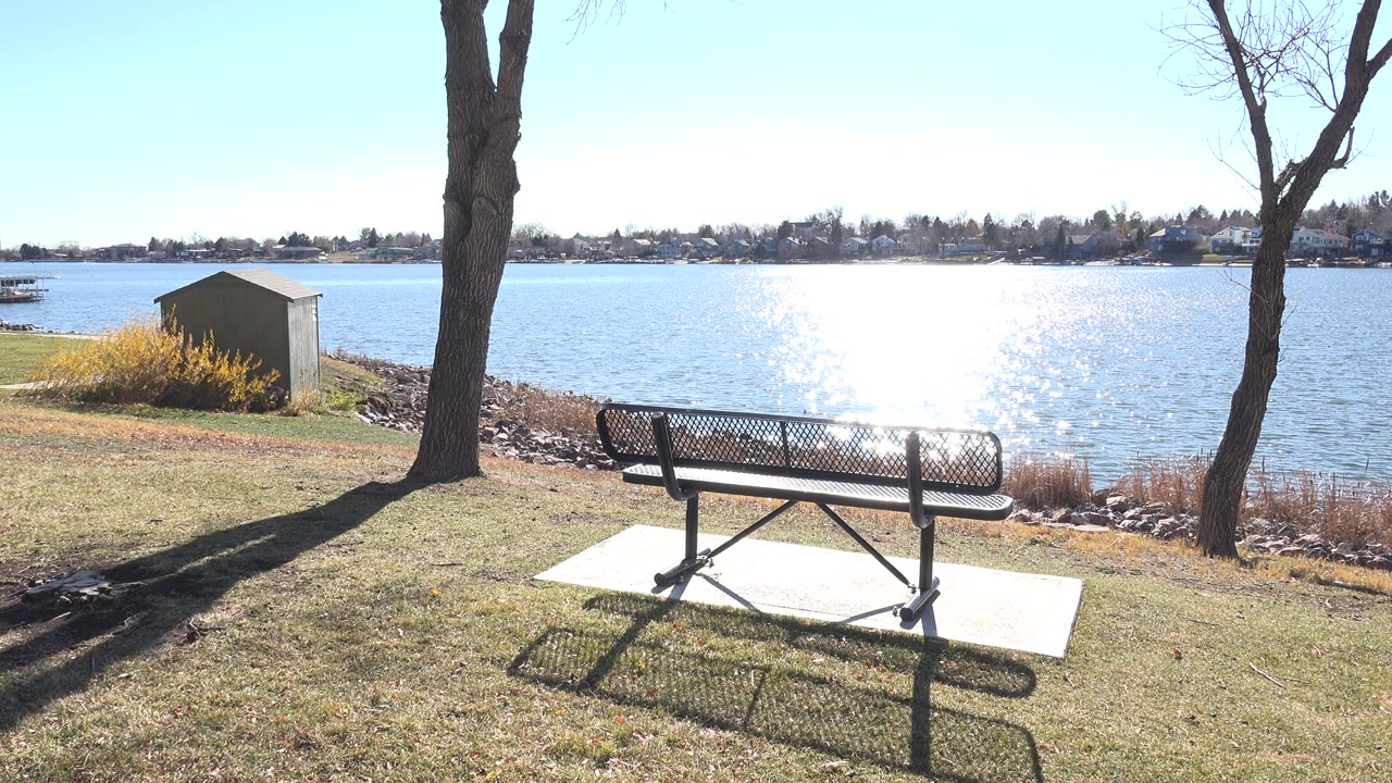 Opening frame at Hidden Lake Colorado with a black metal bench on a concrete pad, bare tree trunks framing the lake, bright sun glare on the water, and no visible people.