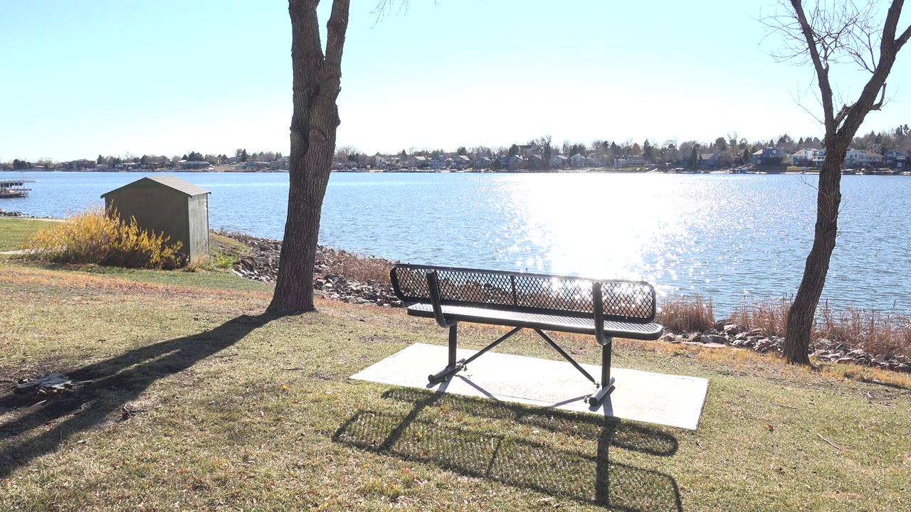Second frame keeps the bench centered near the lower edge at Hidden Lake Colorado, with the lake glare spreading behind it and no visible people.