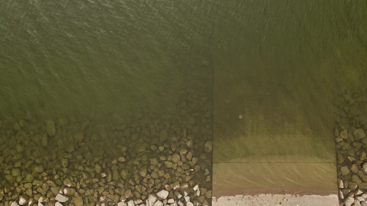 Top-down view of green lake water meeting a rocky shoreline and a narrow ramp in Jefferson County, Colorado, with no visible people.