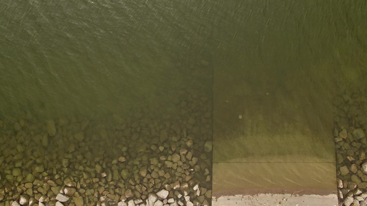 Green ripples pass over shallow lake water beside shoreline rocks and a pale ramp in Jefferson County, Colorado, with no visible people.