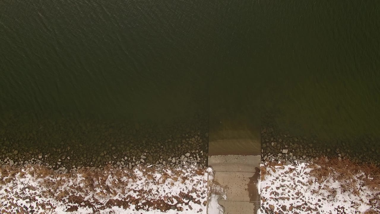Aerial view of green water meeting a snowy rocky bank with a concrete segment at the edge.