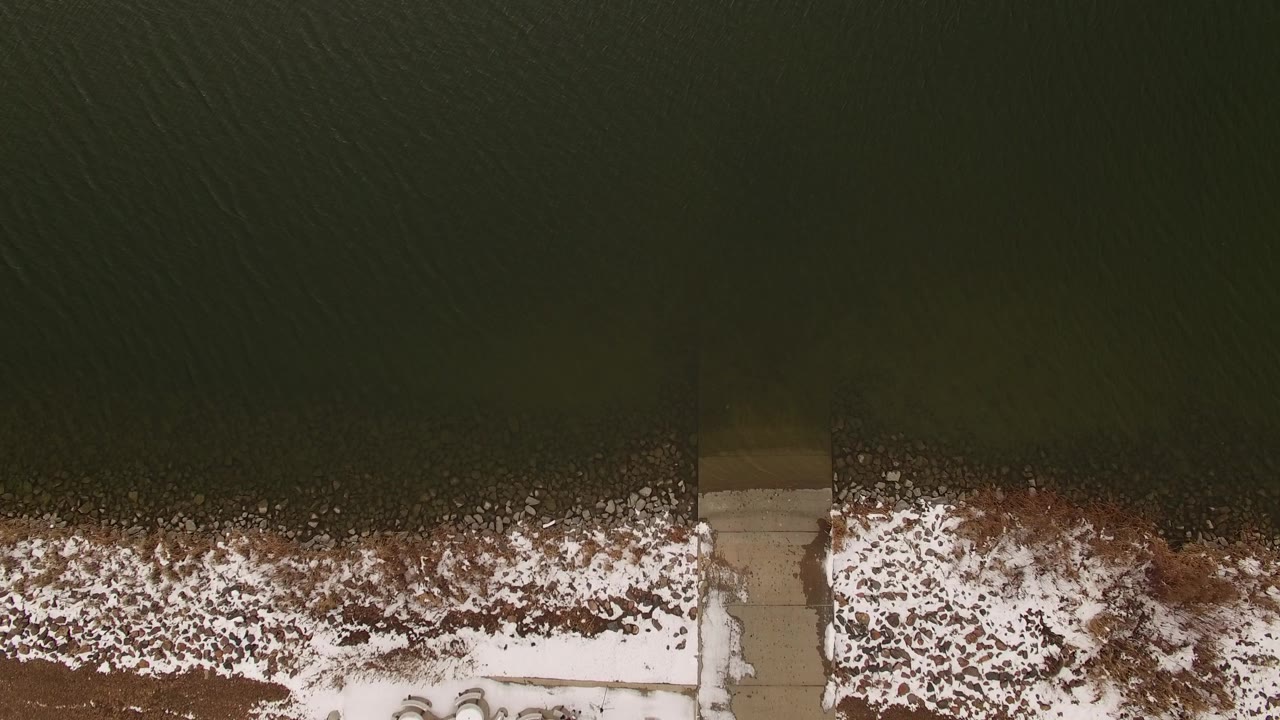 Top-down frame from Jefferson County, Colorado showing dark water above a patchy snowy shoreline with a narrow concrete dock near the center and no visible people.