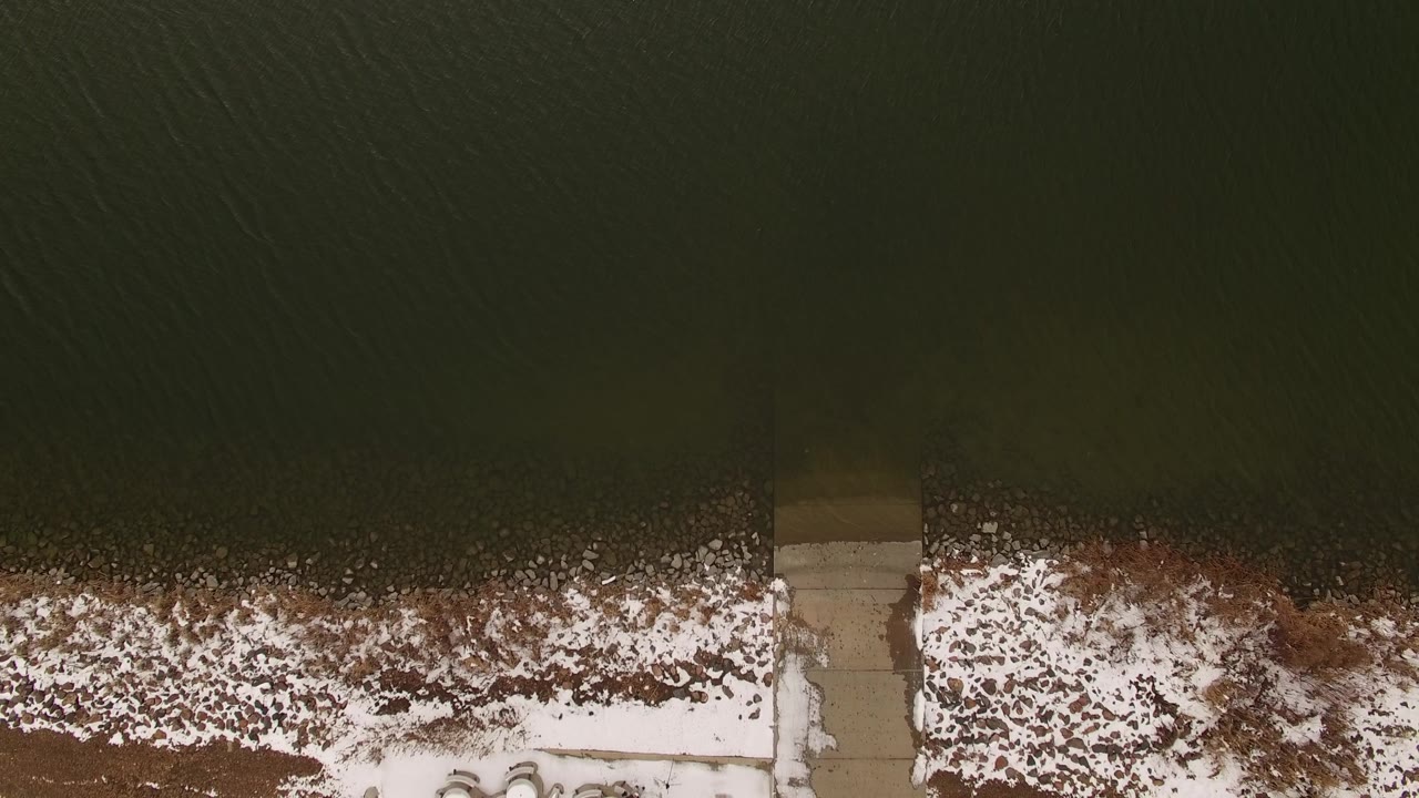 Aerial overhead frame in Jefferson County, Colorado with dark water filling the upper area, snow along the bank, a dock crossing the shoreline, and no visible people.
