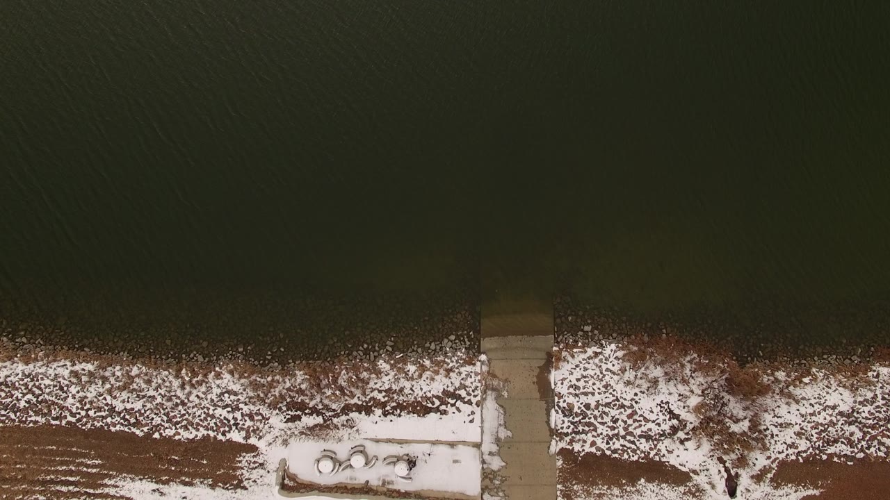 Overhead view in Jefferson, Colorado of dark water meeting a snow-lined shore with a straight dock near the lower edge and no visible people.