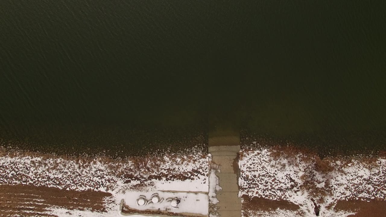 Aerial frame in Jefferson, Colorado showing a pale dock crossing the snowy shoreline below dark green water, with no visible people.