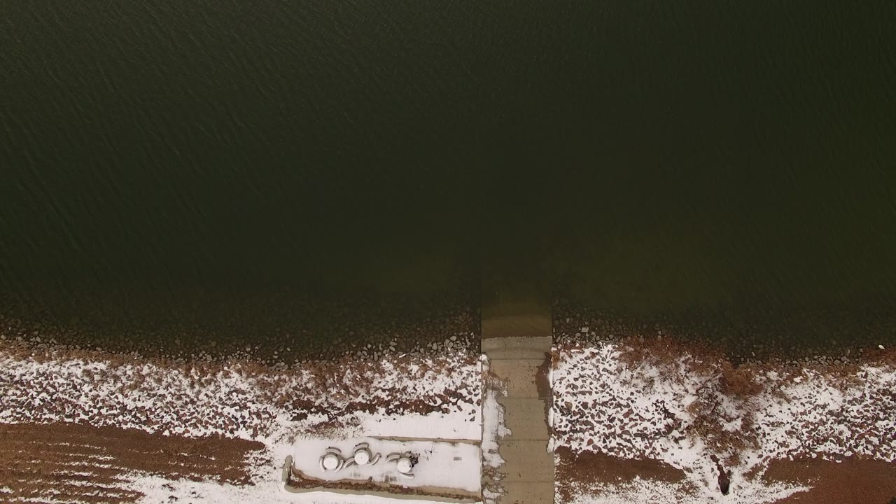 Top-down winter shore with water, snow, and leafless brush.