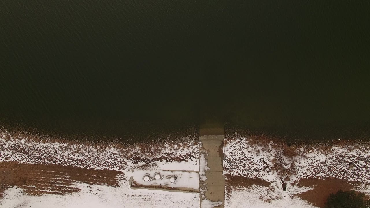 Overhead view of dark water beside a snowy shoreline and concrete path