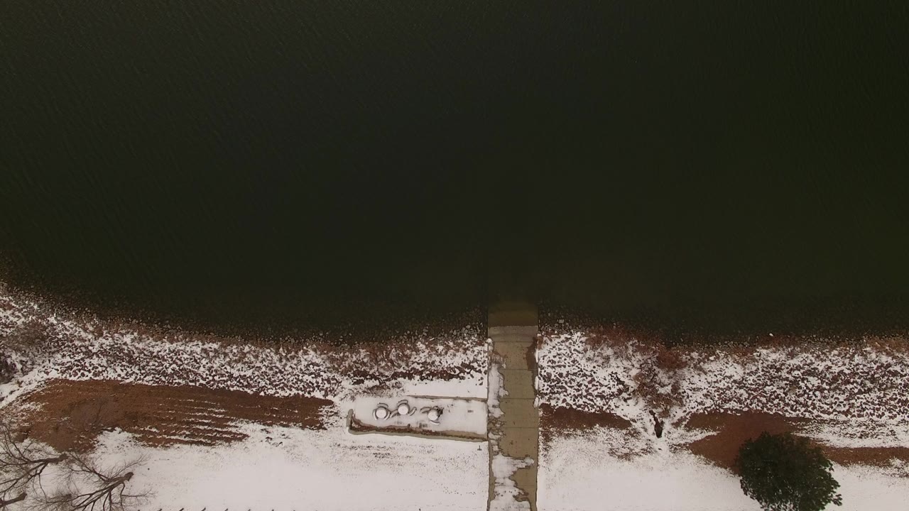 Overhead snowy shoreline with dark water and a straight dock or walkway.