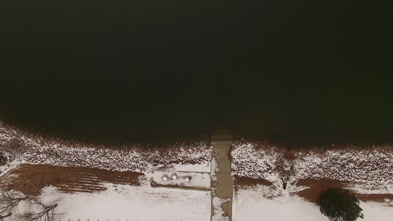 Top-down Jefferson County, Colorado frame with dark water above, a thin snow line at the shore, brown ground cover, a small dock or platform area, and no visible people.