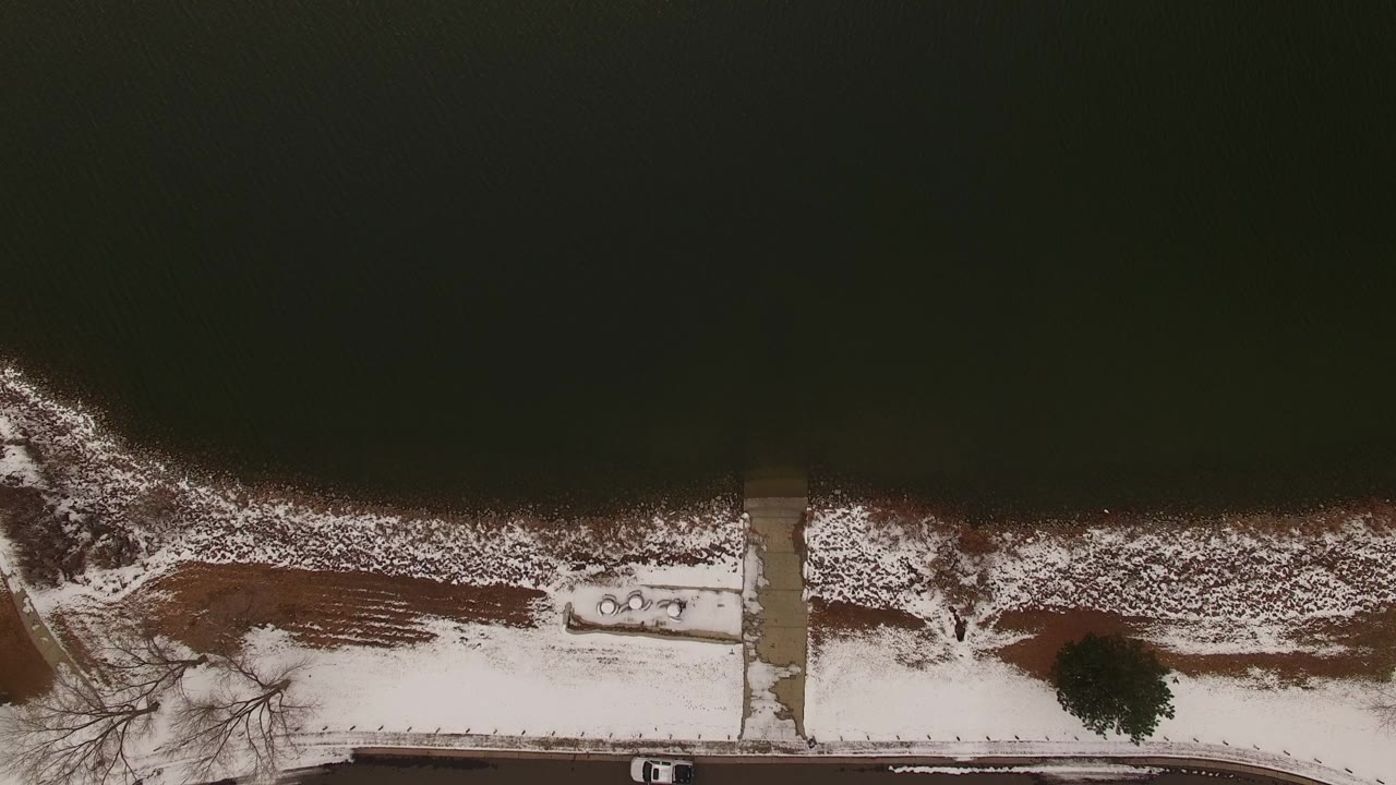 Overhead view in Jefferson County, Colorado showing dark water above a snow-lined shore, a road, a bare tree, and no visible people at the start of the clip.