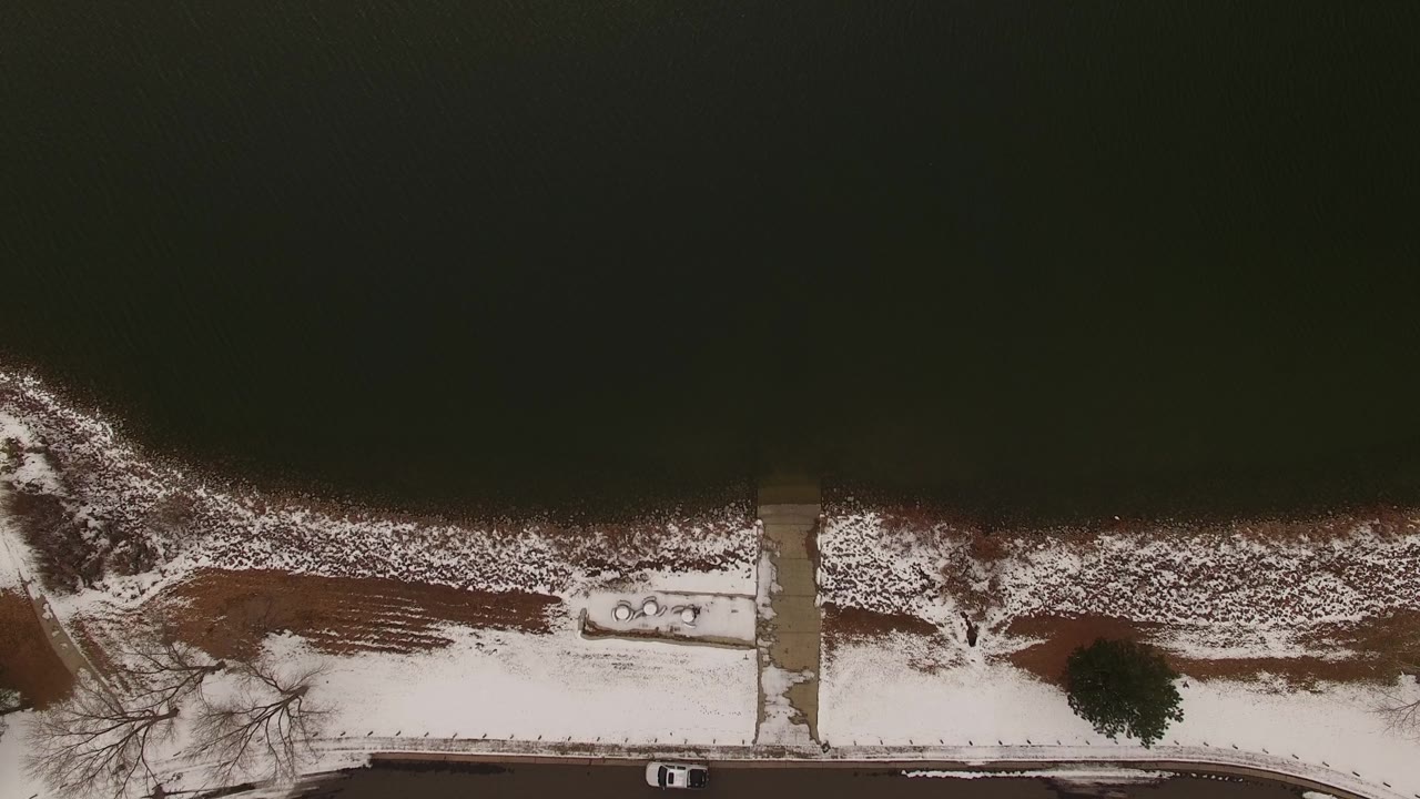Overhead winter shoreline with water, snow patches, path, and car