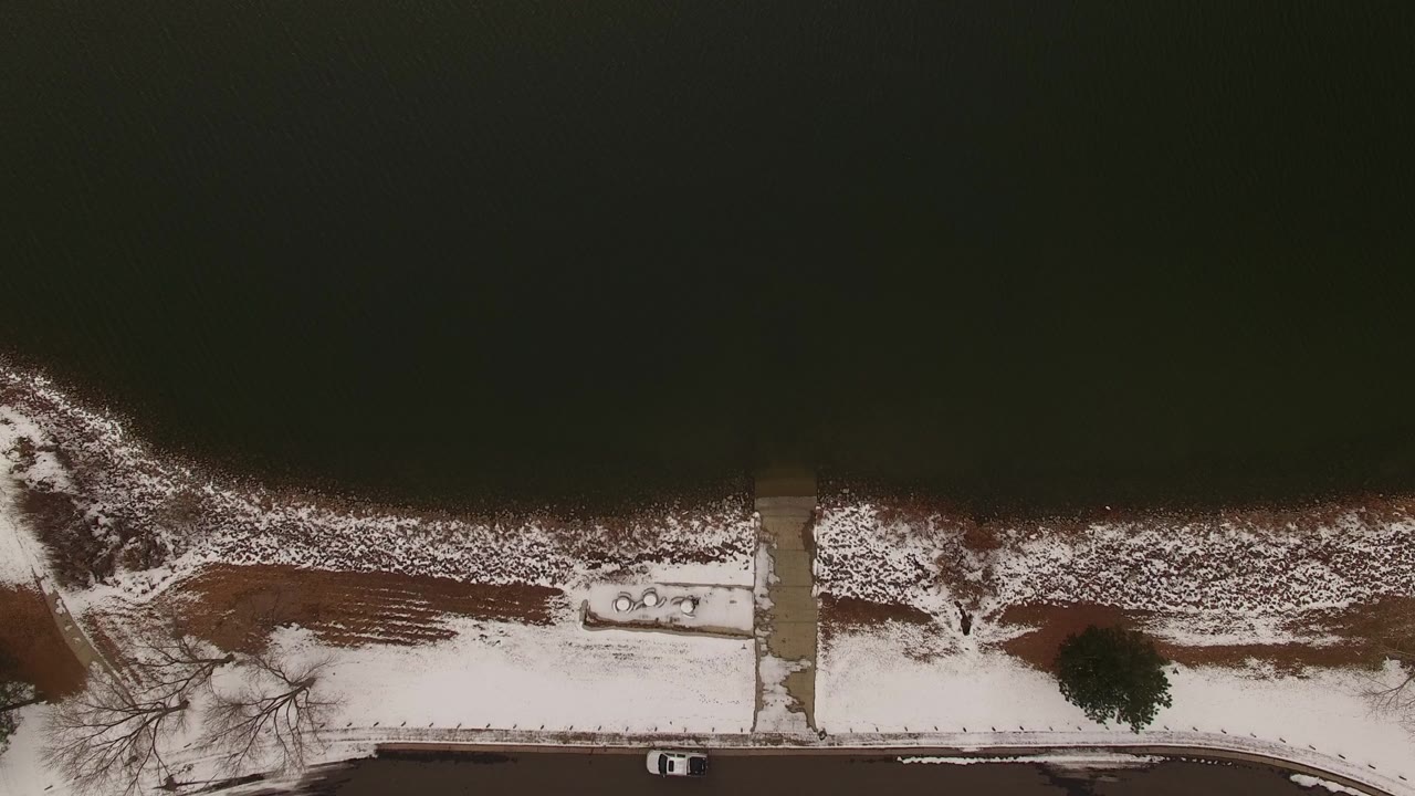 Top-down Jefferson County, Colorado aerial frame showing dark lake water, patchy snow along the shoreline, a sidewalk path, a road, a parked vehicle, and no visible people.