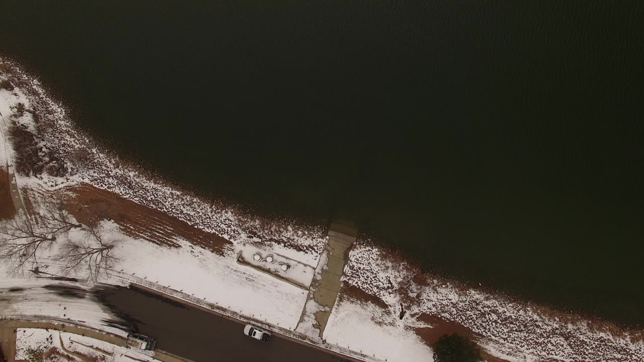 Diagonal aerial view of lake shoreline, road, houses, and snow.