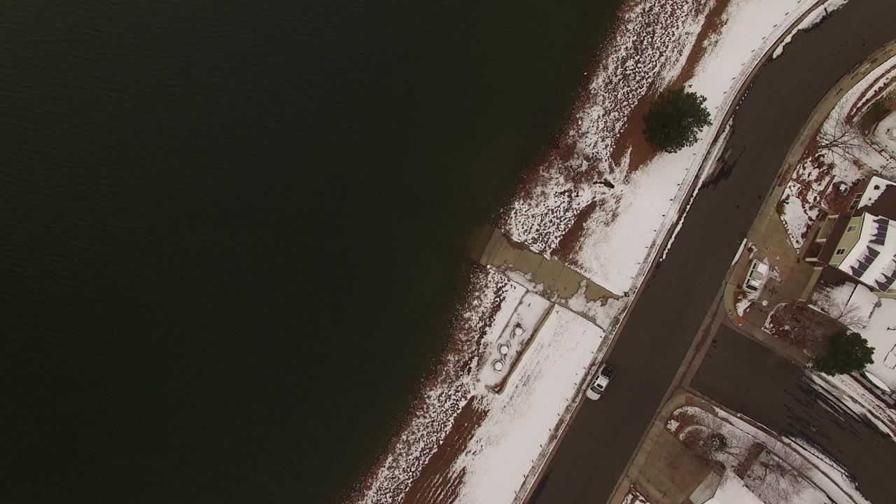 Top-down snowy shoreline beside dark water and a curved road.
