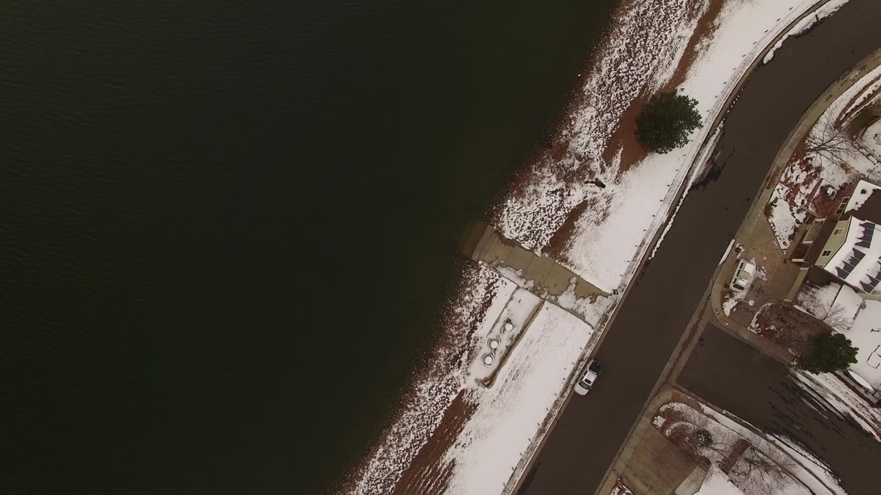 Aerial frame over Jefferson County, Colorado with dark water at left, snow along the shoreline, a road and sidewalks at right, small residential buildings, cars, and no visible people.