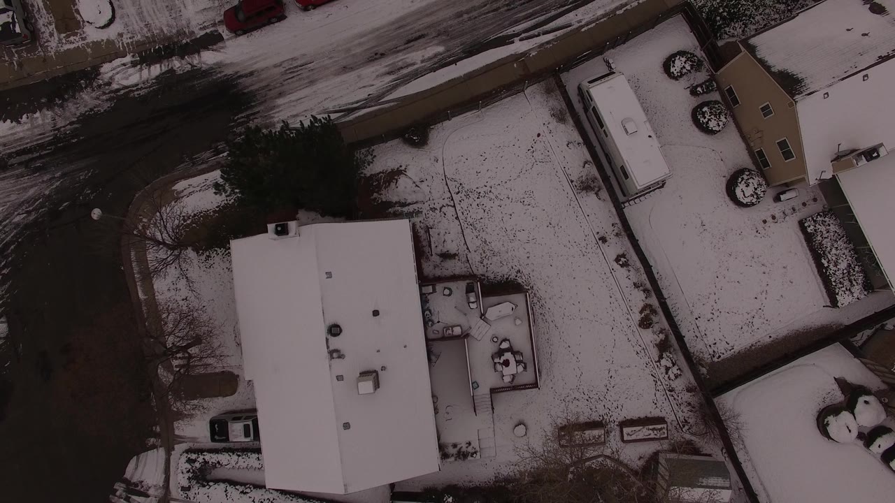 Aerial view over Jefferson County, Colorado residential lots with a large white roof, narrow driveways, parked vehicles, snow patches, and no visible people.