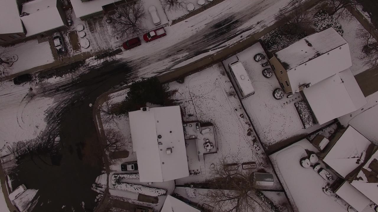 Overhead snowy neighborhood with houses, roads, and parked cars.