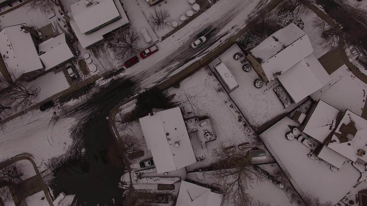 Aerial frame over Jefferson County, Colorado showing snow-covered rooftops, a curving dark road, bare trees, parked vehicles, and no visible people.