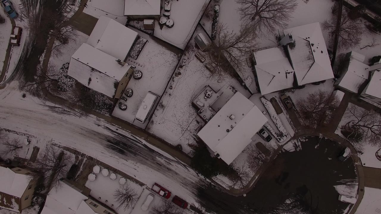 A near-vertical aerial view over Jefferson County, Colorado shows snow-covered homes beside a dark curved road, parked cars, bare trees, and no visible people.