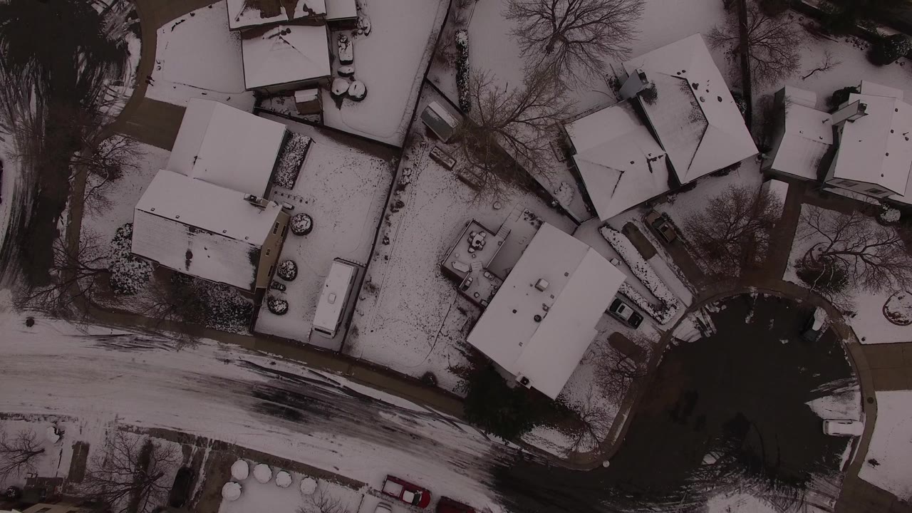 Overhead frame of Jefferson County, Colorado homes with snow-covered roofs, bare trees, a dark road, parked vehicles, and no visible people.