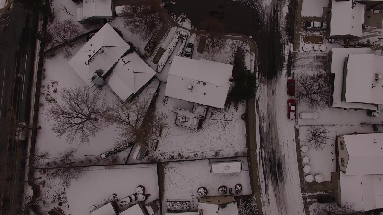 Overhead snowy neighborhood with homes, trees, parked cars, and a road
