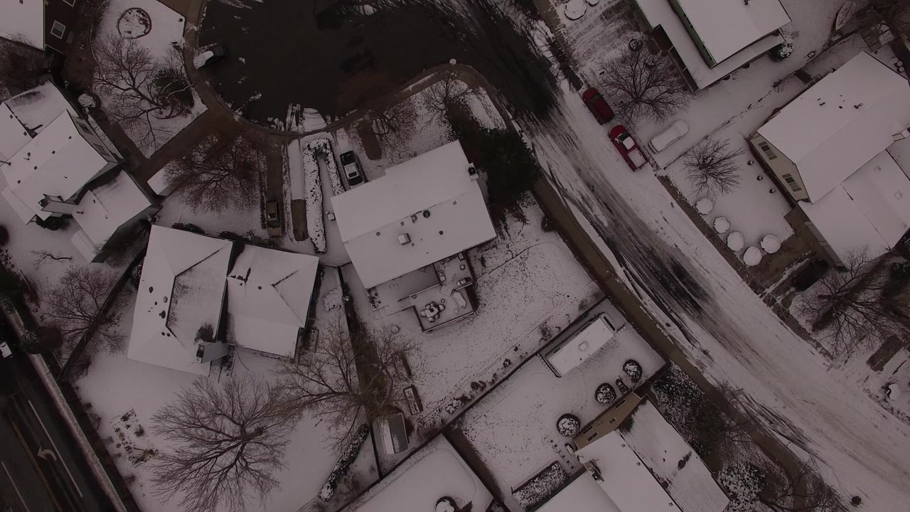 Overhead snowy neighborhood with houses, trees, roads, and parked cars.