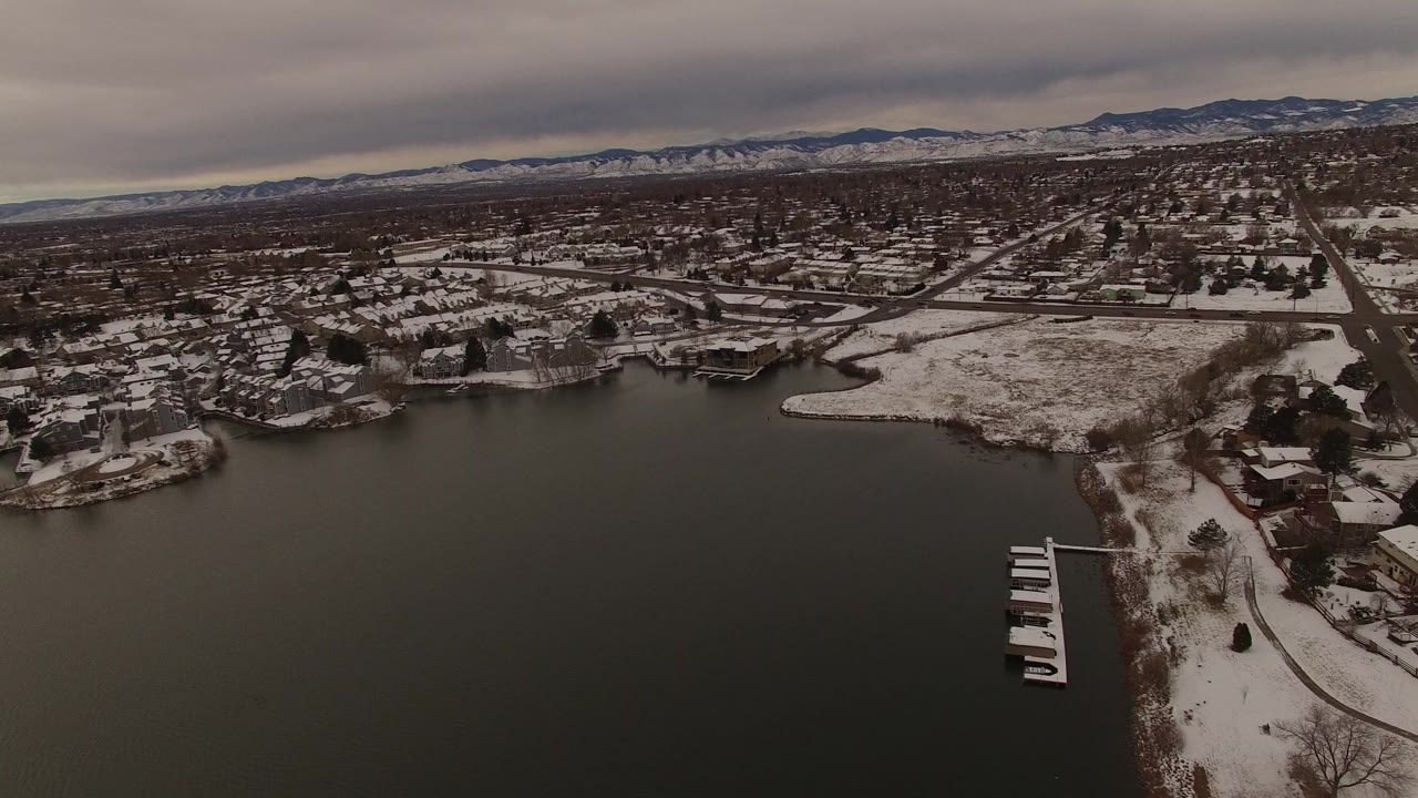 Opening aerial frame over a Jefferson County, Colorado lake, with dark water in front, snowy houses and roads beyond, mountains on the horizon, and no visible people.