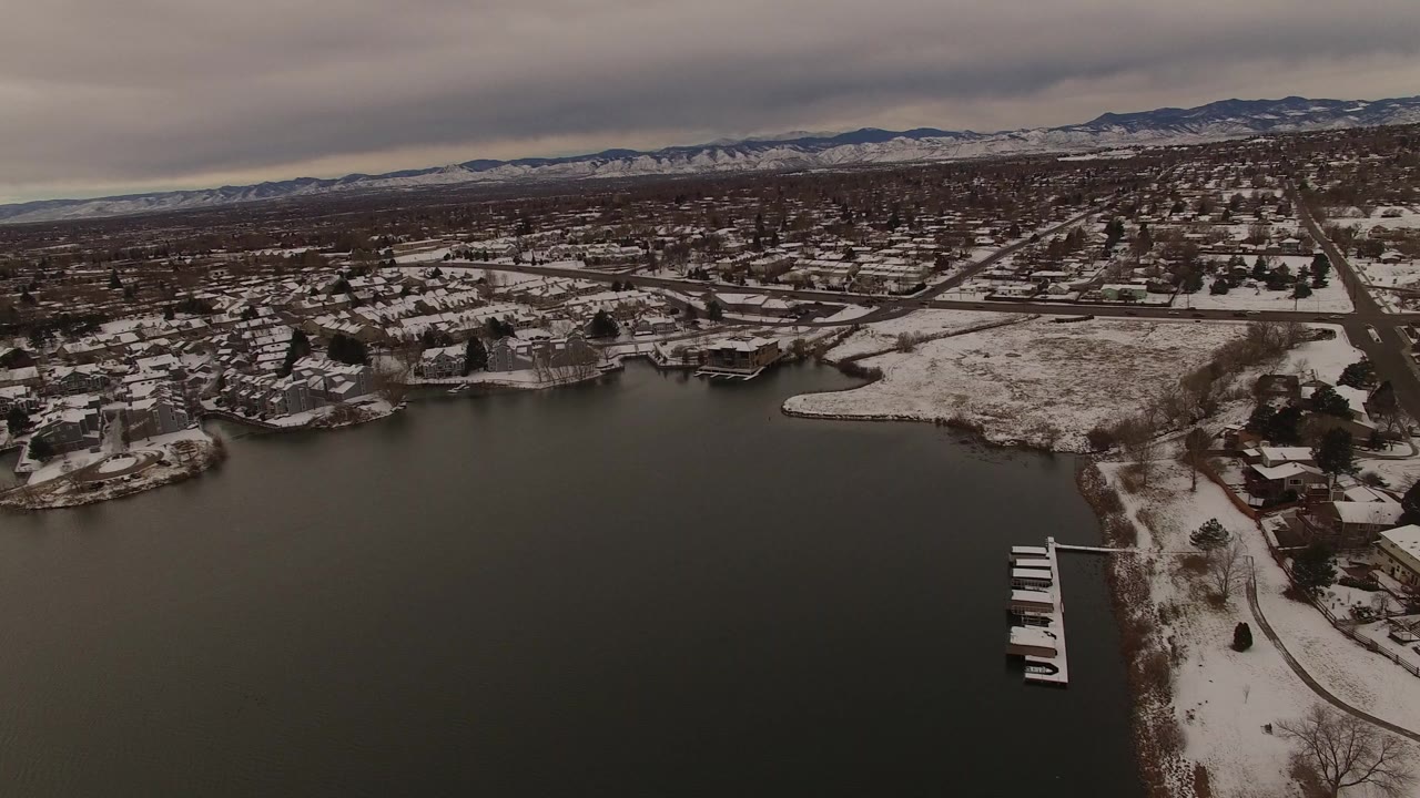 Aerial frame in Jefferson County, Colorado showing the lake foreground, a curved snow-edged shoreline, residential streets, small docks, distant mountains, and no visible people.