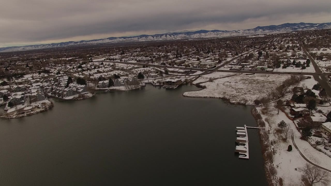Drone-like aerial over a winter lake with snow-dusted houses, piers, roads, and mountains beyond.