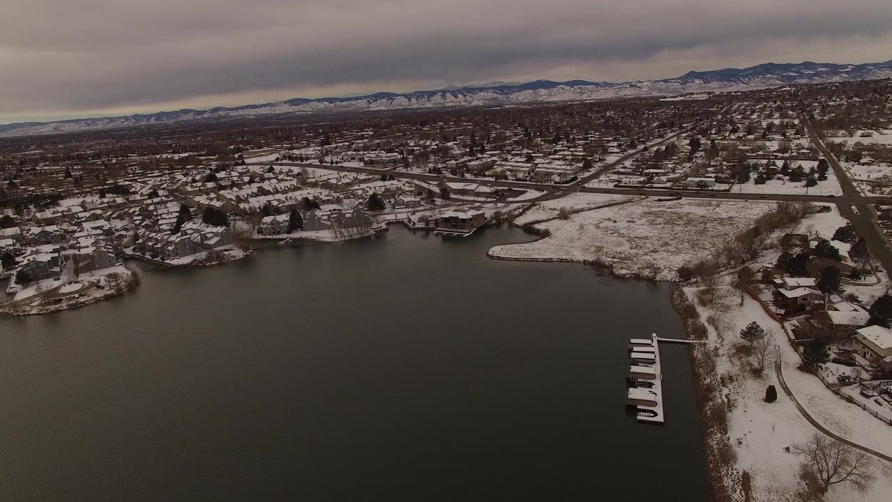 Aerial view in Jefferson, Colorado looking over a dark bay toward snow covered shoreline homes, roads, docks, and distant mountains under gray clouds, with no visible people.