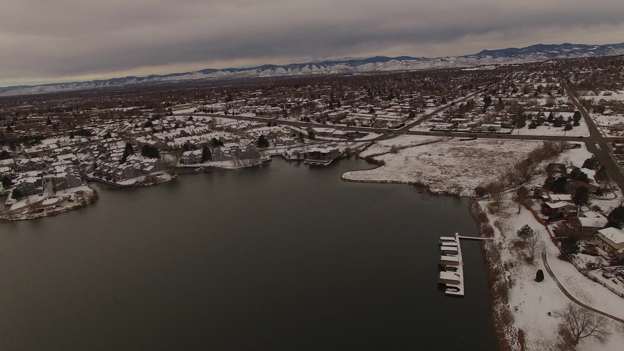 Aerial frame in Jefferson, Colorado showing the bay across the lower frame, snowy homes and roads across the shore, small docks at right, and a mountain horizon, with no visible people.
