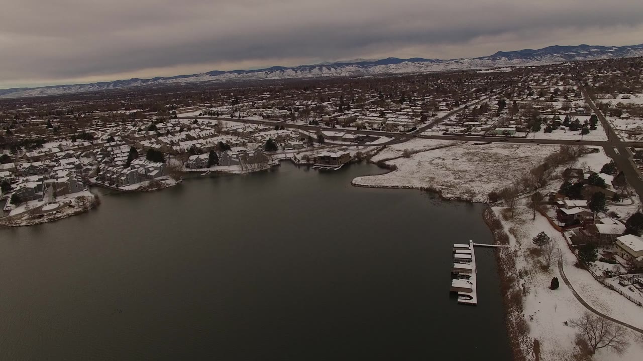 Aerial winter view over bay water toward shoreline houses and distant mountains.