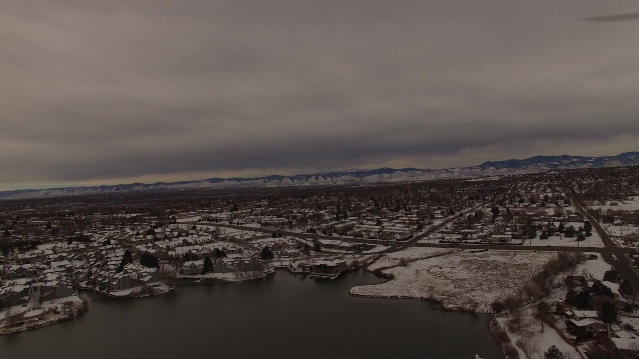 High aerial frame over Jefferson County, Colorado, showing a lake edge, clustered houses, gridded roads, winter trees, distant mountain ridges, and no visible people.