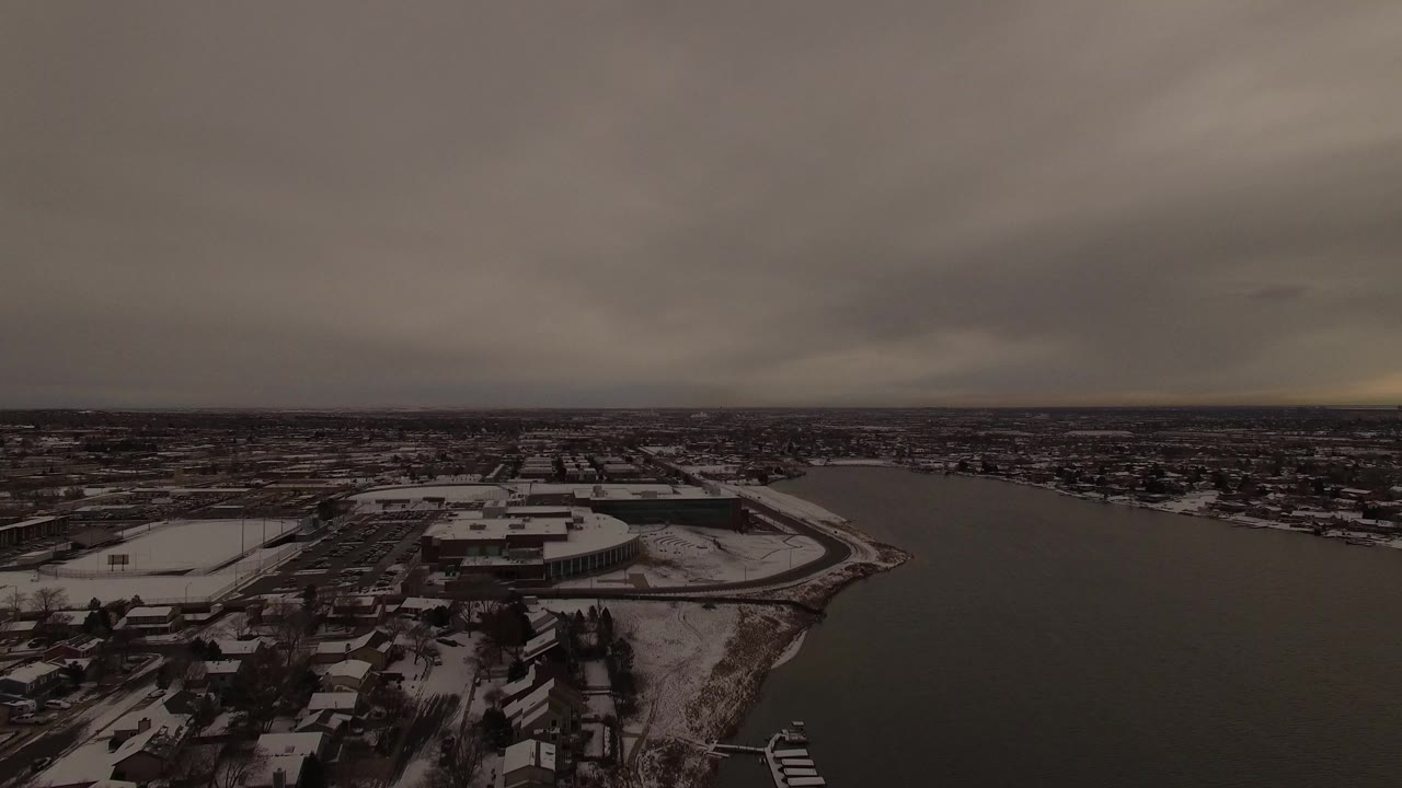 Aerial view of snowy homes beside a dark body of water under gray clouds.