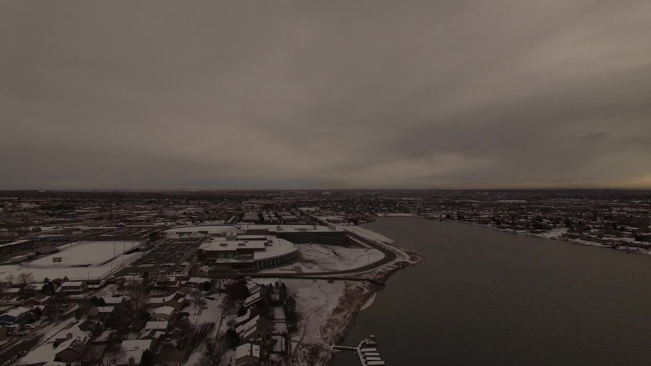 High aerial frame in Jefferson County, Colorado with a snowy shoreline neighborhood, dark water through the center, distant houses, and no visible people.
