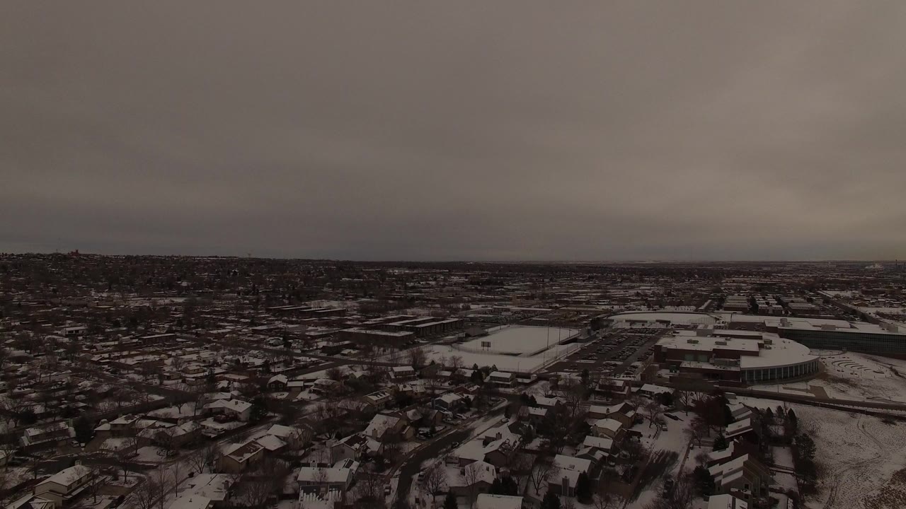 Aerial view over Jefferson County, Colorado showing snow-dusted homes, roads, large flat-roof buildings, and a low overcast horizon with no visible people.