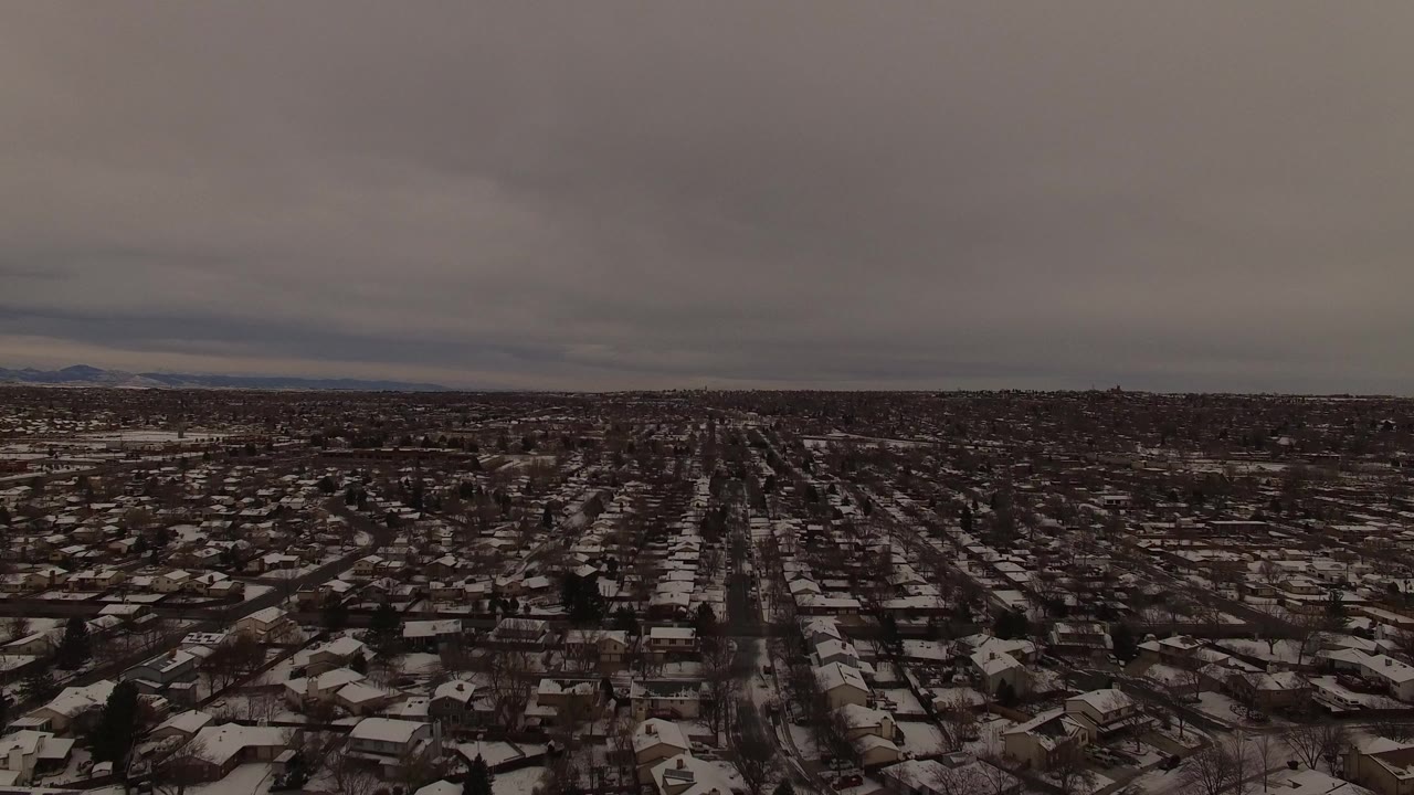Wide aerial view over a snow-dusted Jefferson County, Colorado suburb with gridded streets, rooftops, distant mountains, and no visible people.