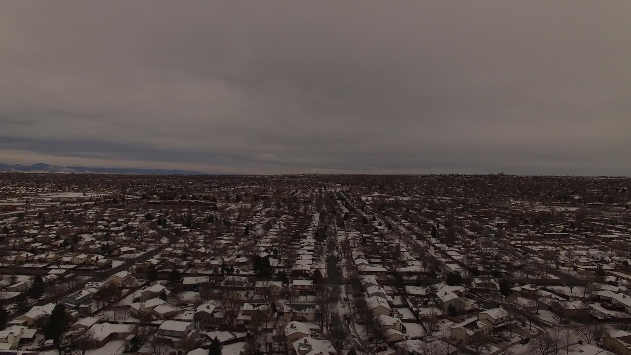 Aerial frame of a Jefferson County, Colorado neighborhood with snow on roofs and yards, a straight street corridor, gray clouds, and no visible people.