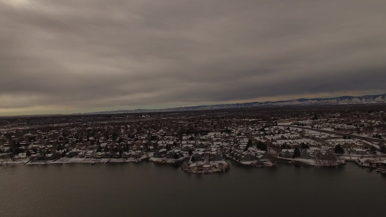 Opening aerial frame over Jefferson County, Colorado, with dark lake water in front, snow-covered residential shoreline beyond it, distant hills, heavy gray clouds, and no visible people.