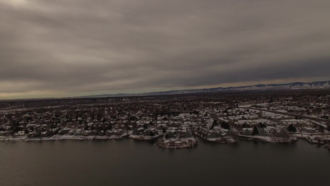 Early frame from Jefferson County, Colorado, showing a broad snowy neighborhood along the lake edge, dark water below, distant low hills, a flat horizon, and no visible people.