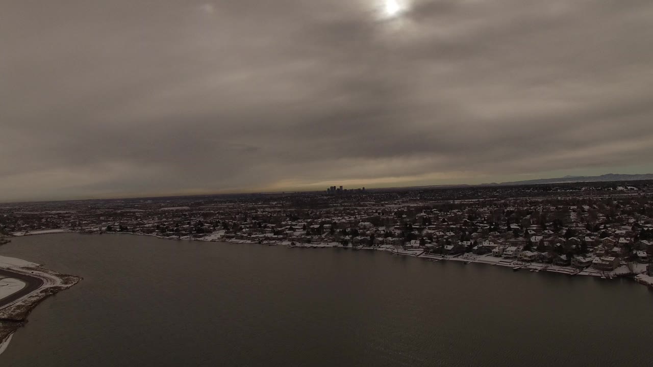 Aerial view over a dark lake toward snowy lakeside homes and a distant skyline.