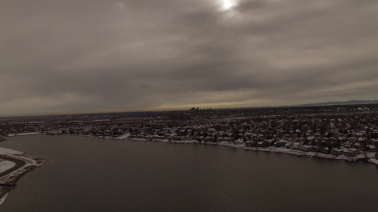 Jefferson County aerial view holds across the lake with snowy homes stretching along the shoreline, a small skyline on the horizon, and no visible people.
