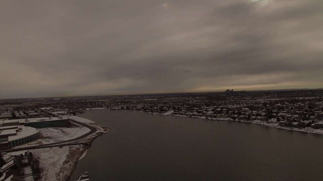 Wide aerial view of a dark lake, snowy shore buildings, and heavy clouds.