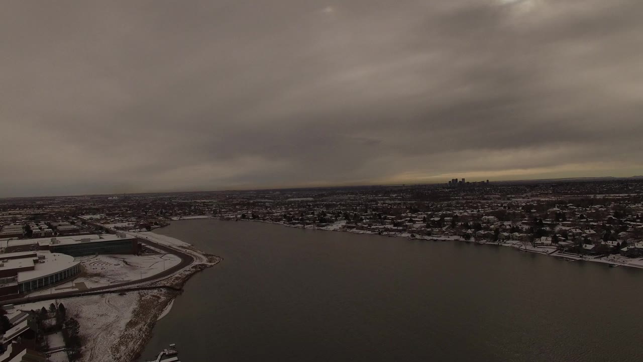 Aerial view over the Jefferson County, Colorado lake with the snow-edged shore slightly shifted in frame, low lakeside structures, muted water, heavy clouds, and no visible people.