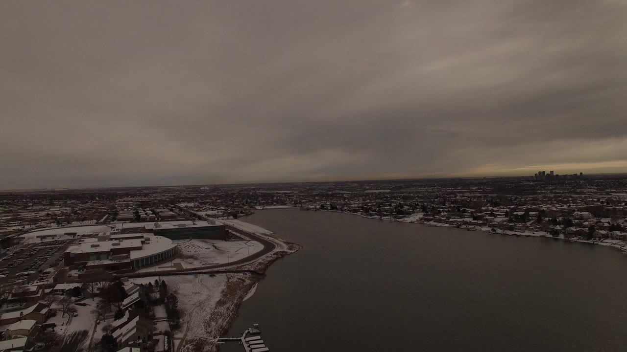 Opening aerial frame over Jefferson County, Colorado showing a dark lake, snow-covered shoreline buildings, distant town blocks, heavy gray sky, and no visible people.