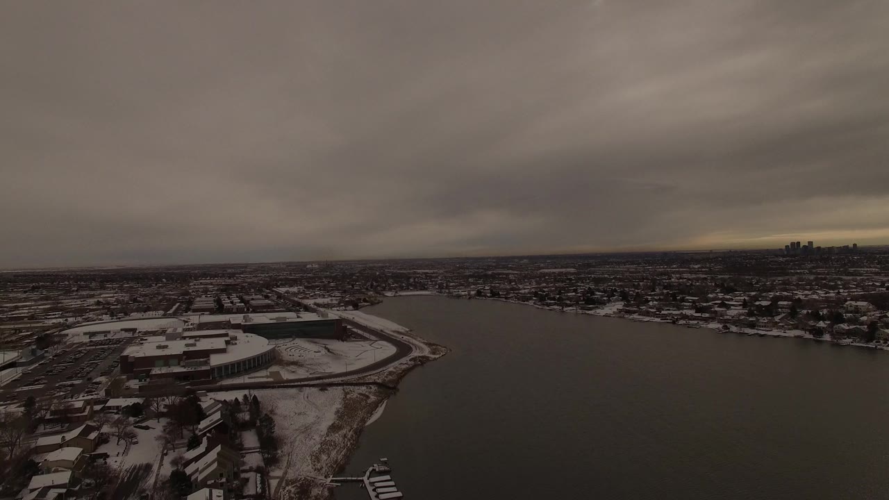 Snowy shoreline and dark water seen from above with a town in the distance.
