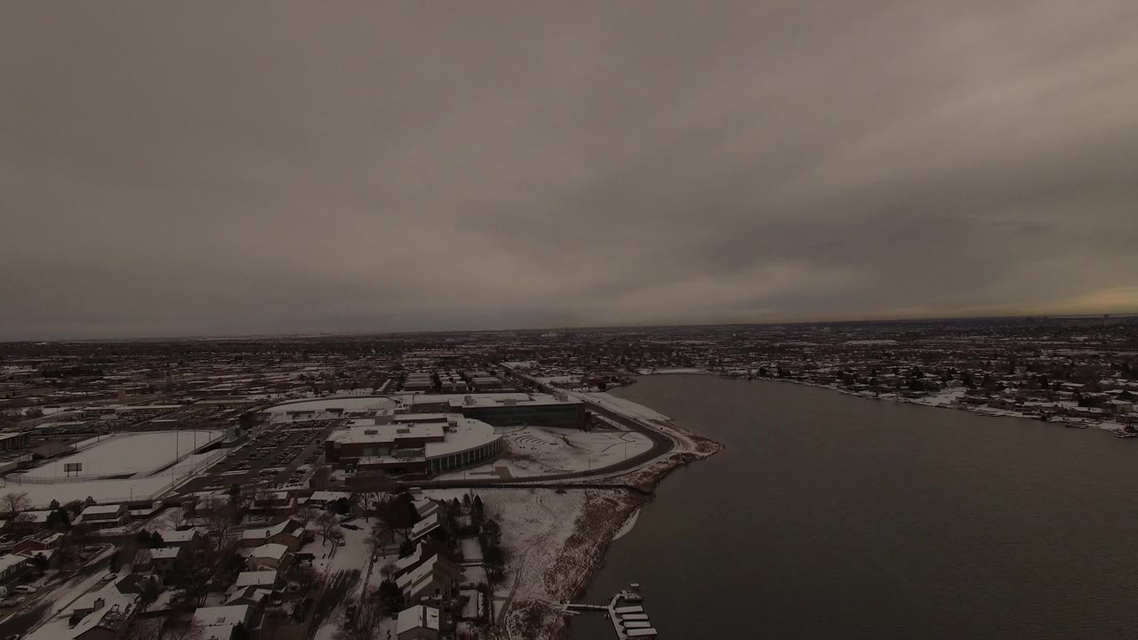 High aerial frame in Jefferson County, Colorado showing a dark lake bordered by snow-covered homes, shoreline docks, streets, and distant low buildings beneath heavy cloud cover, with no visible people.