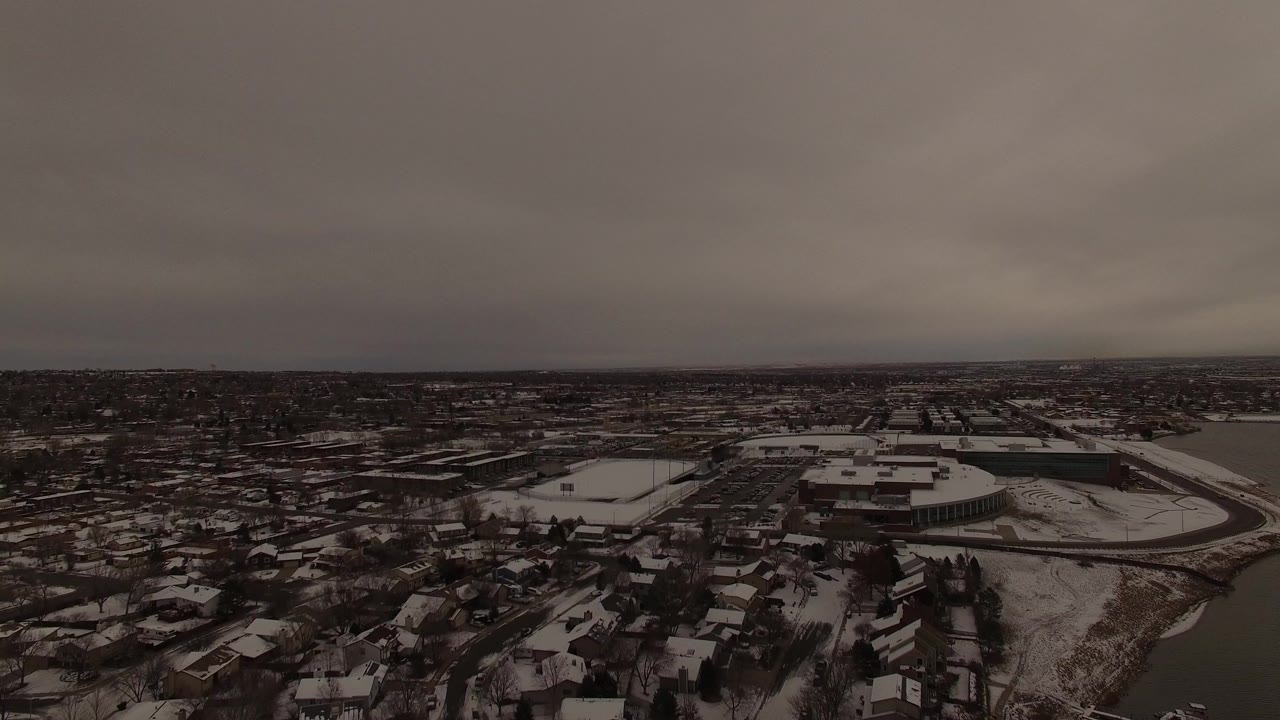 Early aerial view over Jefferson County, Colorado, showing snow on residential rooftops, bare trees, larger buildings, and a dark overcast horizon with no visible people.