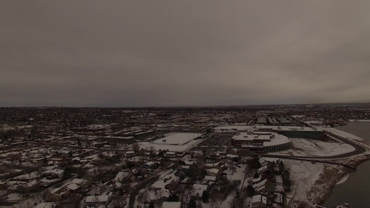 Aerial frame over Jefferson County, Colorado, with a curving waterway or pond edge near snowy houses, large buildings, bare trees, and no visible people.