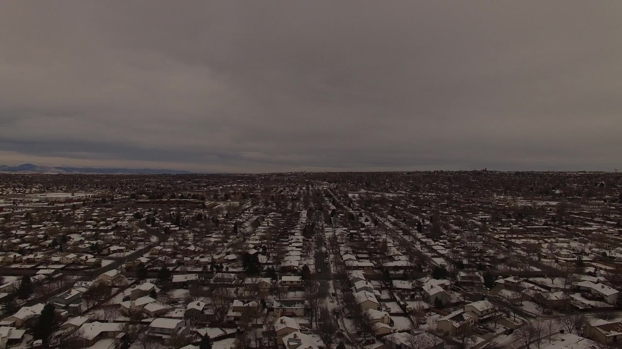 Aerial frame over Jefferson County, Colorado showing long straight residential streets, snow on roofs and lawns, and a dark winter horizon with no visible people.